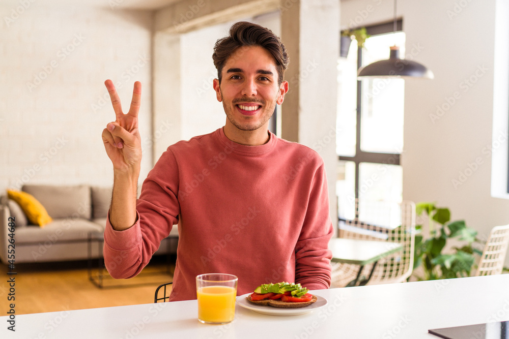 Young mixed race man having breakfast in his kitchen showing number two with fingers. Young mixed race man having breakfast in his kitchen showing number two with fingers.
