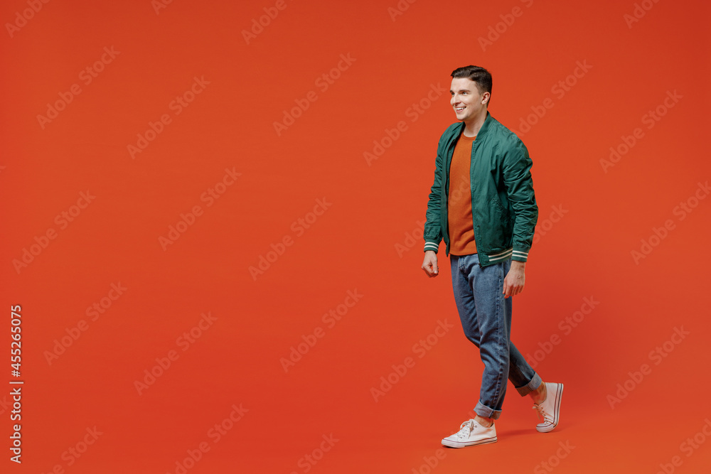 Fototapeta premium Full size body length side view profile vivid happy young brunet man 20s wears red t-shirt green jacket go move isolated on plain orange background studio portrait. People emotions lifestyle concept.