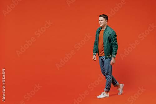 Full size body length side view profile vivid happy young brunet man 20s wears red t-shirt green jacket go move isolated on plain orange background studio portrait. People emotions lifestyle concept.