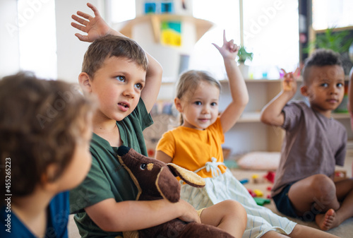 Group of small nursery school children sitting on floor indoors in classroom  raising hands.