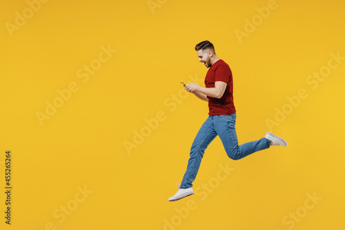 Full length profile side view happy young man in red t-shirt casual clothes jump high using mobile cell phone isolated on plain yellow color wall background studio portrait. People lifestyle concept.