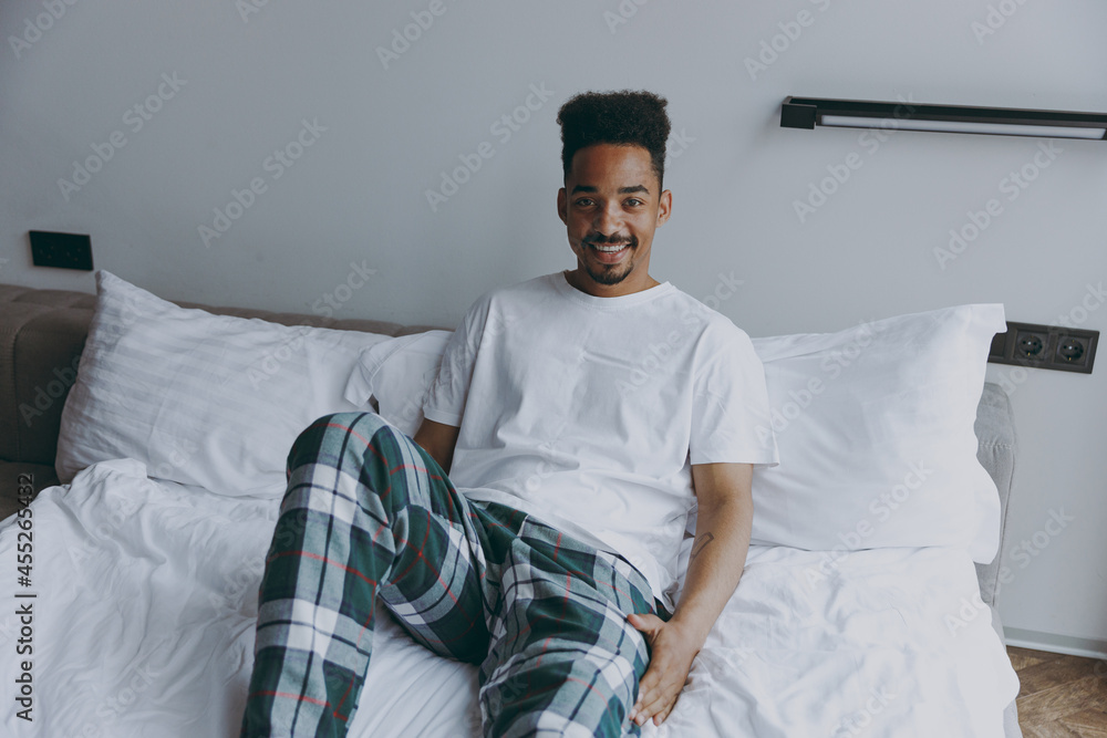 Young happy smiling african american man wearing pj bottoms lying in bed rest relax spend time in bedroom lounge home in own room house wake up dream be lost in reverie good day. Real estate concept Young happy smiling african american man wearing pj bottoms lying in bed rest relax spend time in bedroom lounge home in own room house wake up dream be lost in reverie good day. Real estate concept