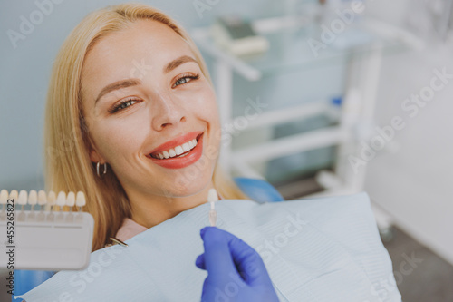 Close up young smiling woman choosing veneers enamel color palette with stomatologist compare teeth shade sit at dentist office chair indoor light modern cabinet Healthcare enamel whitening treatment. photo