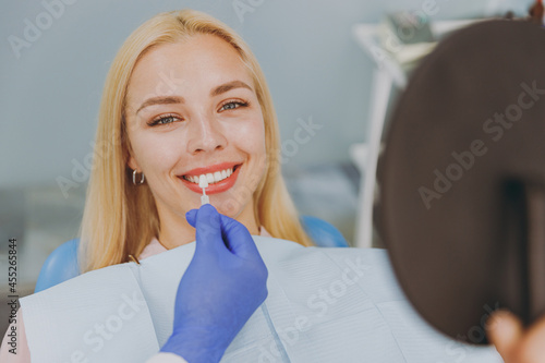 Принти на полотні Young smiling calm happy woman choose veneers enamel color compare teeth shade with stomatologist look at result mirror sit at dentist office chair indoor modern cabinet
