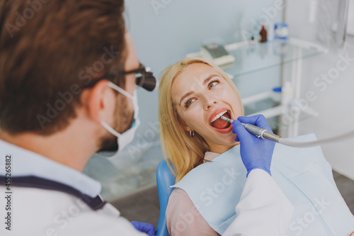 Young man doctor in dentist binocular loupe mask drills tooth of patient caucasian woman with caries decay sit at dentist office chair indoor cabinet near stomatologist Healthcare enamel treatment