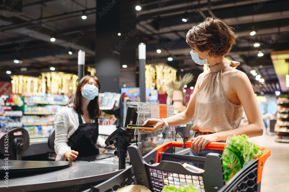 Young woman 20s in casual clothes mask shopping at supermaket with grocery cart put mobile phone to modern bank payment terminal process acquire credit payments at checkout near cashier inside store