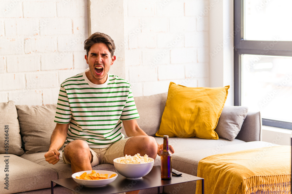Young mixed race man eating popcorns sitting on the sofa screaming very angry and aggressive. Young mixed race man eating popcorns sitting on the sofa screaming very angry and aggressive.