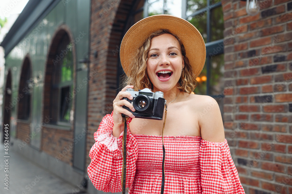Young surprised happy excited woman wear pink dress hold take picture on retro vintage photo camera walk in city standing outdoor near town brick building. People urban summer time lifestyle concept