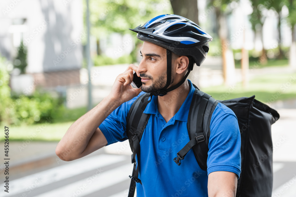 Young muslim deliveryman in safety helmet talking on smartphone outdoors