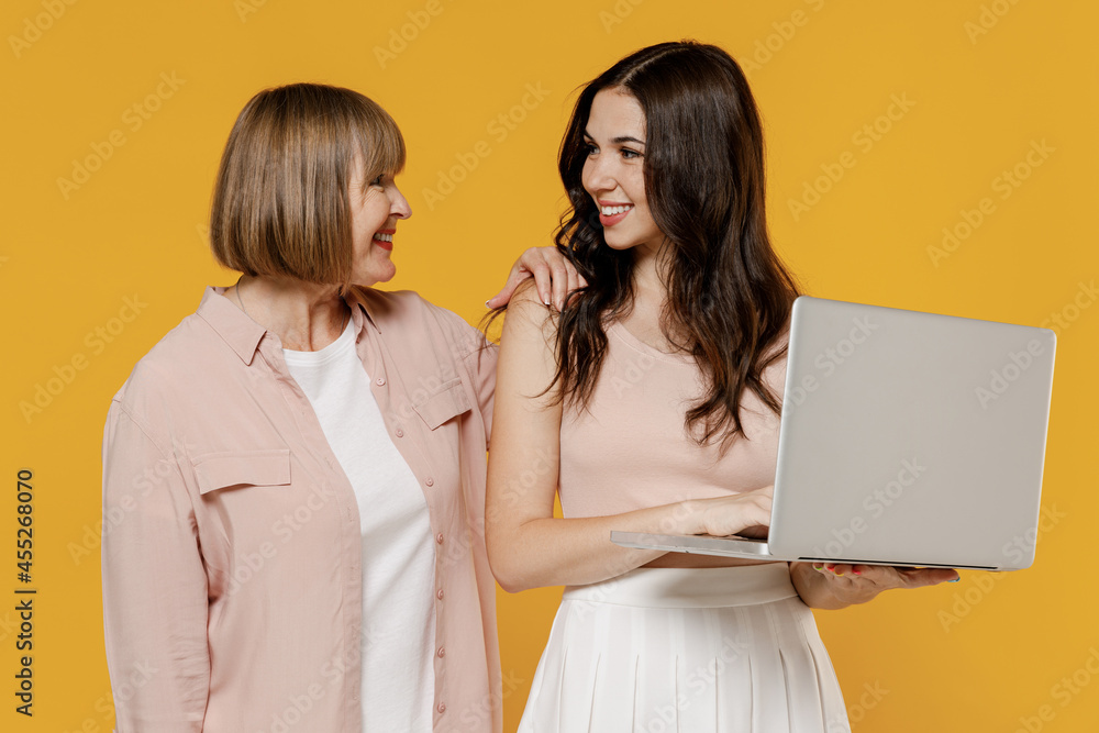 Two young fun daughter mother together couple women wearing casual clothes hold use work on laptop pc computer look to each other isolated on plain yellow background studio Family lifestyle concept. Two young fun daughter mother together couple women wearing casual clothes hold use work on laptop pc computer look to each other isolated on plain yellow background studio Family lifestyle concept.