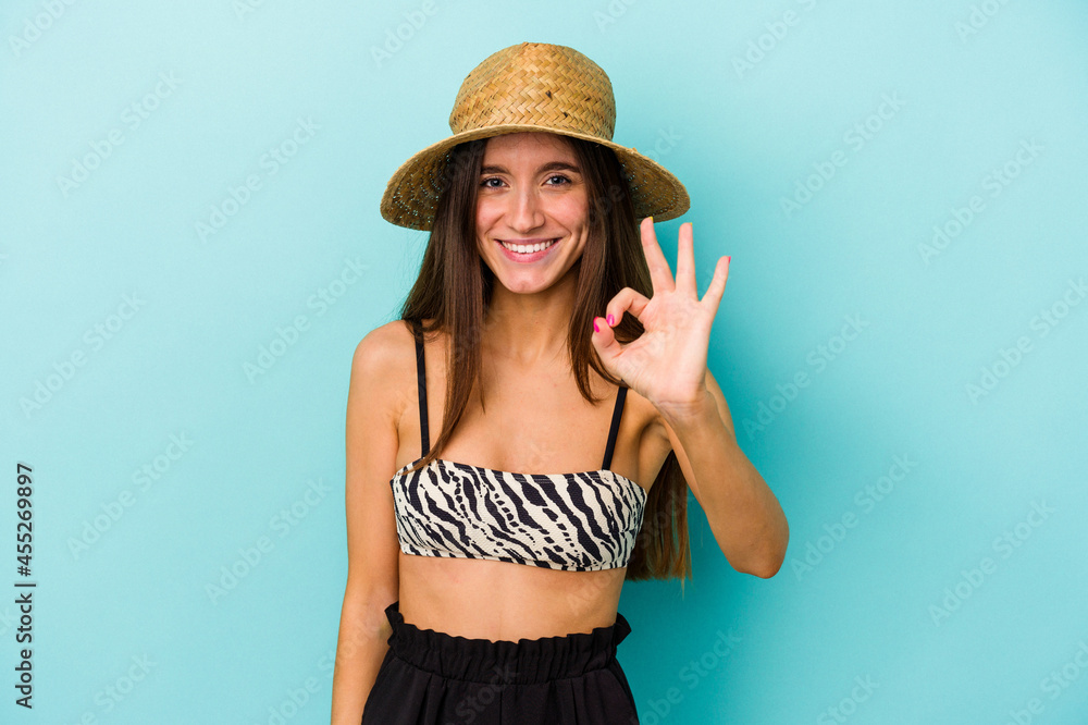 Young caucasian woman going to the beach wearing bikini isolated on blue background cheerful and confident showing ok gesture. Young caucasian woman going to the beach wearing bikini isolated on blue background cheerful and confident showing ok gesture.