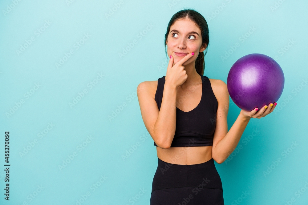 Young caucasian fitness woman holding a ball isolated on blue background looking sideways with doubtful and skeptical expression. Young caucasian fitness woman holding a ball isolated on blue background looking sideways with doubtful and skeptical expression.