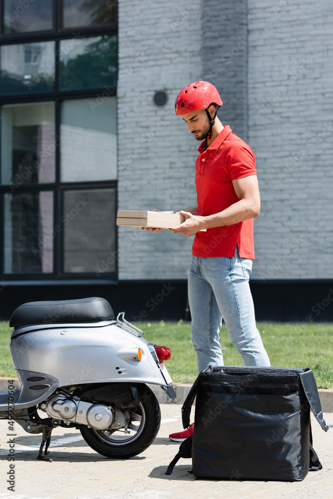 Young muslim courier in helmet holding pizza boxes near scooter and thermo backpack outdoors