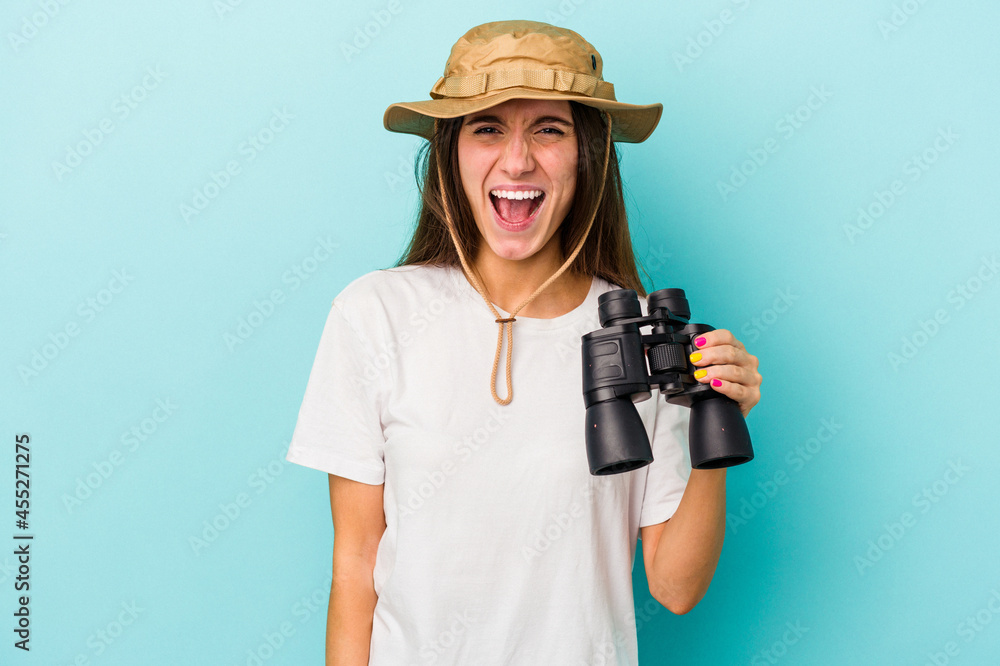 Young caucasian explorer woman holding binoculars isolated on blue background screaming very angry and aggressive. Young caucasian explorer woman holding binoculars isolated on blue background screaming very angry and aggressive.