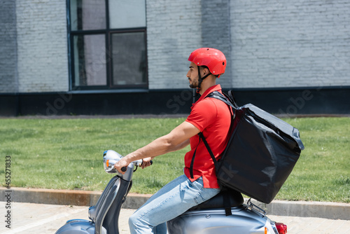 Side view of muslim courier with thermo backpack riding scooter on urban street