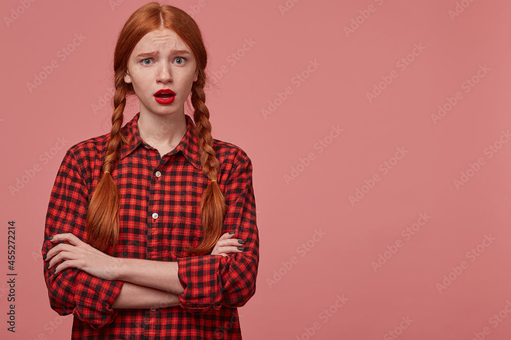 indoor shot of shocked young ginger female keeps her hand crossed, raised her eyebrows with negative facial expression. Isolated over pink background