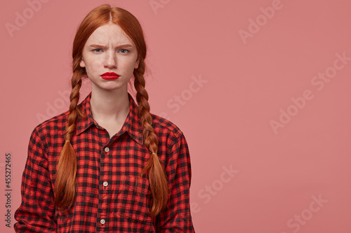 indoor shot of serious and sad young ginger female, pressing her lips and knit eyebrows, starring into camera with confused facial expression. Isolated over pink background with copy space