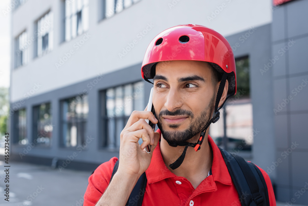 Muslim courier in helmet with backpack talking on smartphone outdoors Muslim courier in helmet with backpack talking on smartphone outdoors