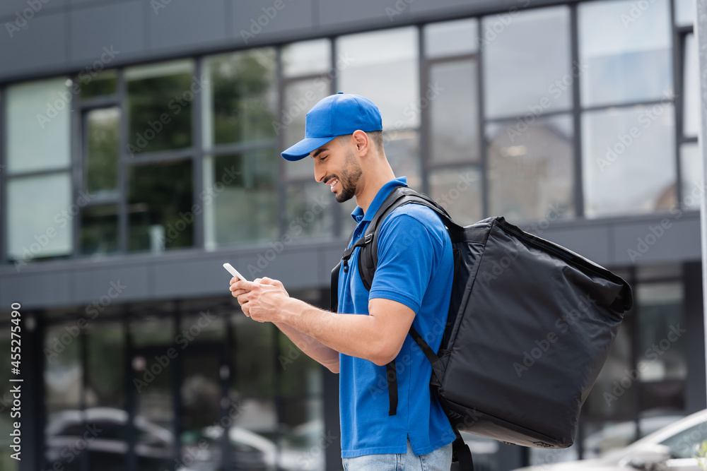 Side view of cheerful arabian courier with thermo backpack using smartphone on urban street Side view of cheerful arabian courier with thermo backpack using smartphone on urban street
