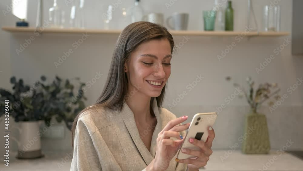 Brunette beautiful woman with phone in hands looking at the screen, smiling. Young woman with device typing and scrolling, gimbal shot, kitchen room with decoration Brunette beautiful woman with phone in hands looking at the screen, smiling. Young woman with device typing and scrolling, gimbal shot, kitchen room with decoration