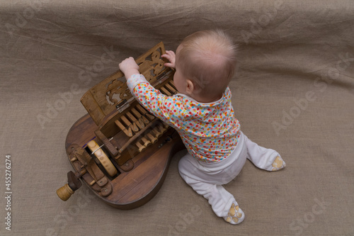 A small nine-month-old child, sitting on his lap, opens the carved cover of the keyboard of an ancient wooden musical instrument - hurdy-gurdy