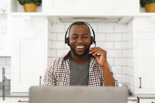 Laughing african american man sitting in kitchen making video call using laptop and headset