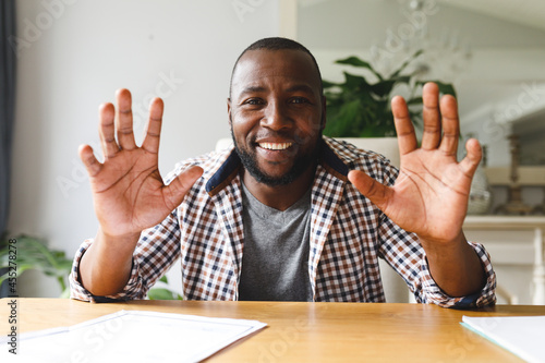 Smiling african american man sitting at table gesturing in dining room, making video call