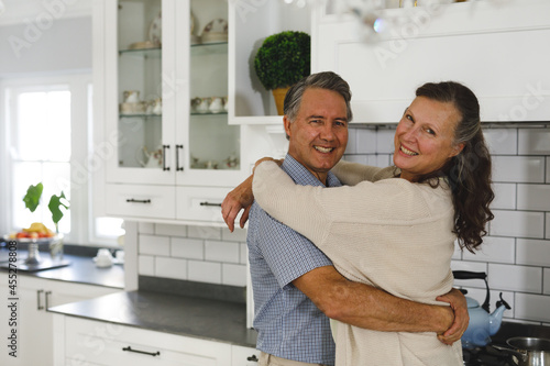 Portrait of happy senior caucasian couple in modern kitchen, embracing, looking to camera