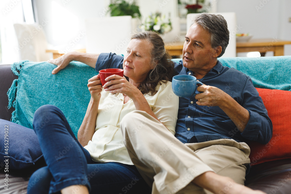 Happy senior caucasian couple in living room sitting on sofa, drinking coffee