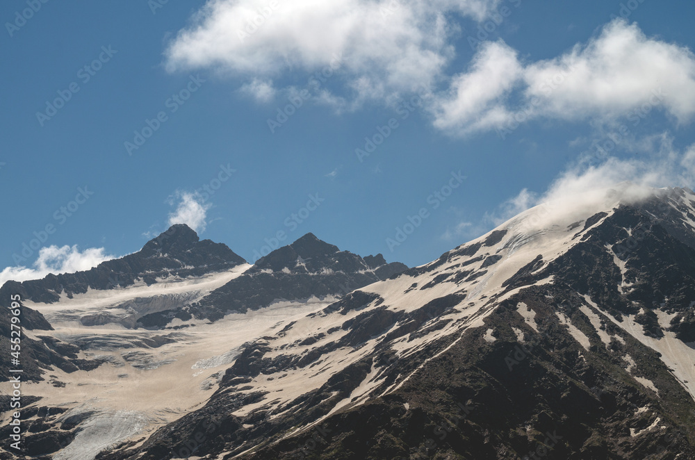 Snowy mountain slope with clouds on the background of a blue sky. The Caucasus mountains in summer.