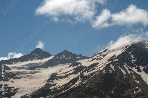 Snowy mountain slope with clouds on the background of a blue sky. The Caucasus mountains in summer.