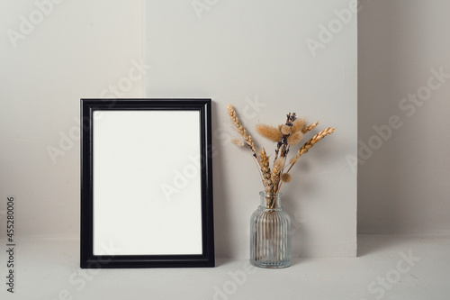 Still life with a black frame. Next to it is a glass vase with dried herbs. Light background. © Алексей Шандалин