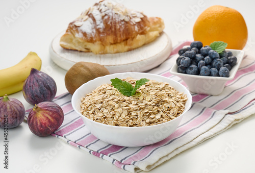 Raw oatmeal in white ceramic plate, blueberries, fig, orange, banana, on white table, breakfast