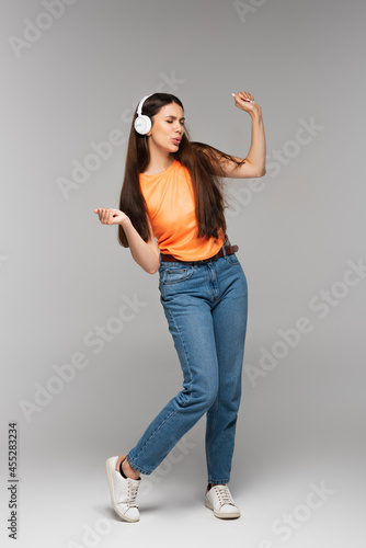 full length of young woman in denim jeans and wireless headphones listening music while dancing on grey