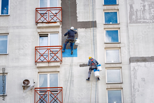  Industrial alpinist at height on rope, plastering wall with trowel. Industrial climber repairing house facade. Rope access job, construction workers repair and restore facade of high building. photo