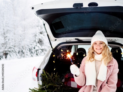 Young woman with christmas sparkler in her hands sitting in the trunk of car. Christmas car trip on the road by winter snow forest. Enjoying nature. Travel concept. Winter season. Christmas holidays