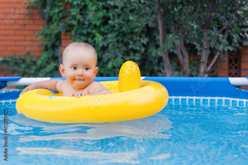 A happy newborn baby of 10-12 months is swimming in a yellow swimming ring. Frame swimming pool in the backyard of the house