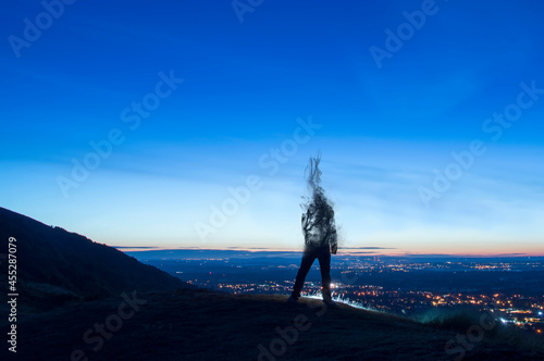 A silhouette of a man disappearing and turning into smoke. Standing on a hill. Looking out on city lights just before sunrise. photo