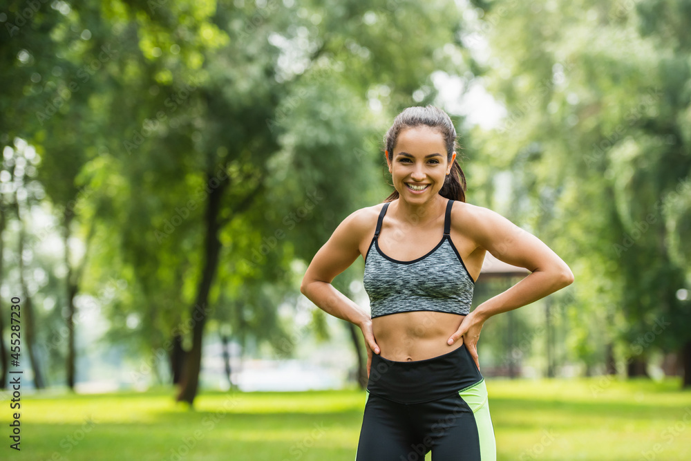 happy athletic woman in crop top resting while standing with hands on hips in green park happy athletic woman in crop top resting while standing with hands on hips in green park