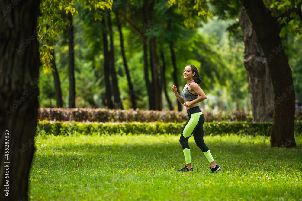 full length of cheerful sportswoman in crop top and leggings jogging on grass in park full length of cheerful sportswoman in crop top and leggings jogging on grass in park