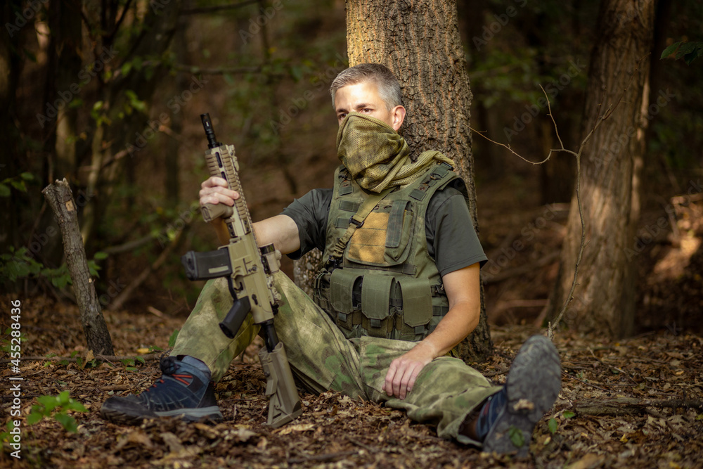 a man is sitting on the ground leaning against a tree with an airsoft assault rifle in military uniform in the forest