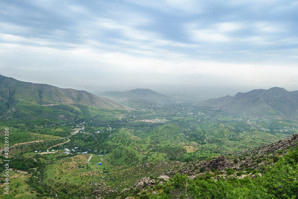 Large green valley surrounded by mountain range. Small village can be seen in distance. Road meanders between hills. Shot in mountains of Zerafshan, Uzbeksistan Large green valley surrounded by mountain range. Small village can be seen in distance. Road meanders between hills. Shot in mountains of Zerafshan, Uzbeksistan
