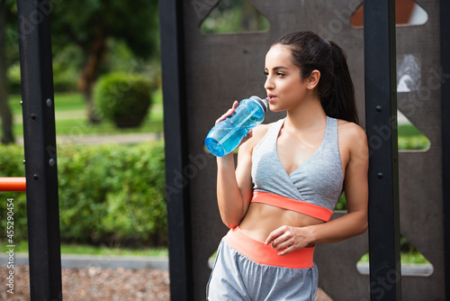 fit woman in sportswear holding sports bottle and drinking water while resting in outdoor gym