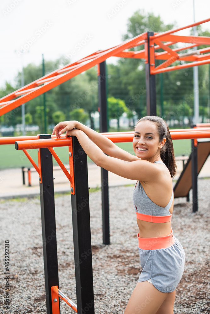 Fototapeta premium cheerful sportswoman smiling while leaning on parallel bars outside