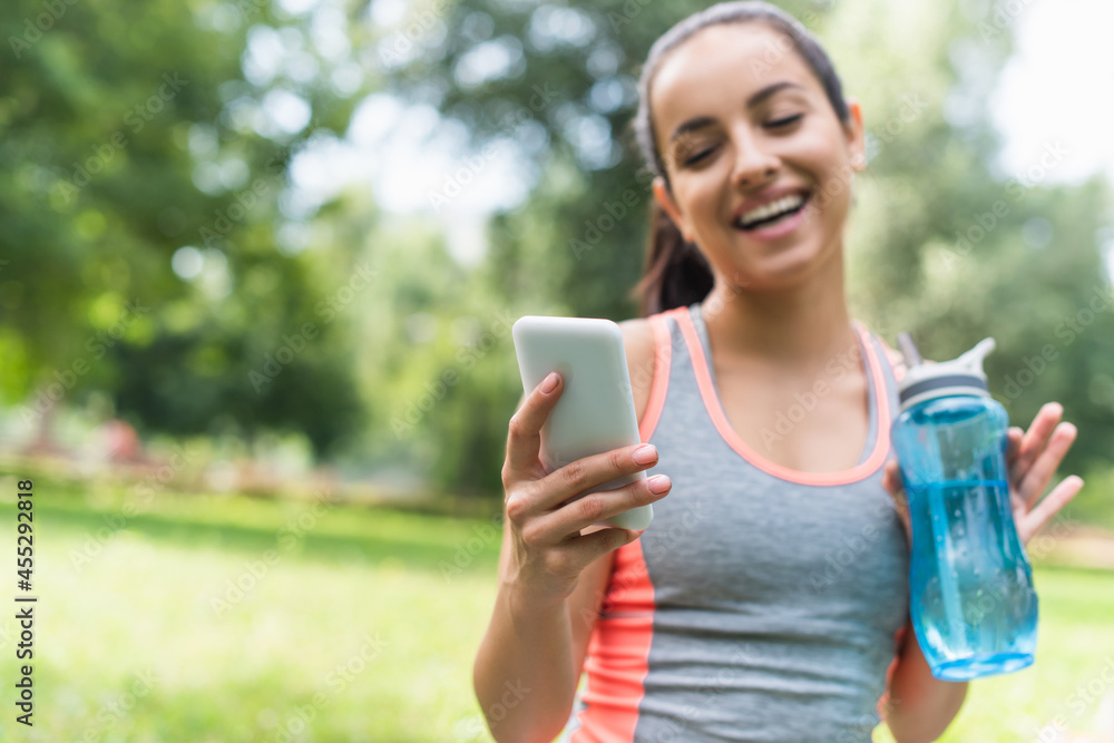 happy and blurred woman holding sports bottle with water and using smartphone in park happy and blurred woman holding sports bottle with water and using smartphone in park
