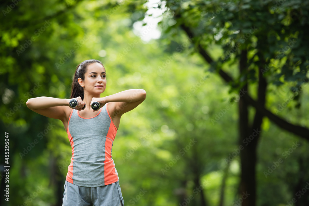 young sportswoman looking away while working out with dumbbells in park young sportswoman looking away while working out with dumbbells in park