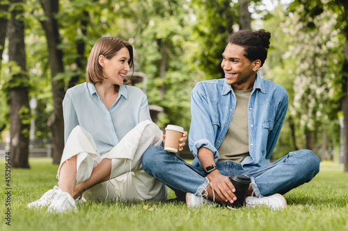 Young mixed-race couple friends boyfriend and girlfriend students spending time together on romantic date sitting on green lawn grass in city park walking outdoors. Love and relationship © InsideCreativeHouse