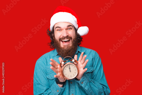 Happy bearded man in santa claus hat holding alarm clock at 12 o clock. photo
