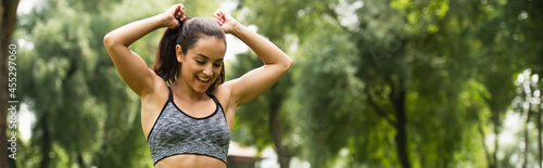 happy athletic woman in crop top adjusting ponytail in green park, banner