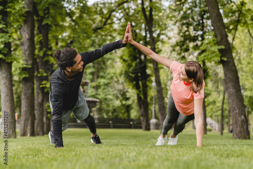 Fit healthy young male and female athletes boyfriend and girlfriend friends couple standing in plank position stretching warming-up before training workout together outdoors in city park.